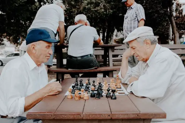 two older men playing chest in the park at a picnic table which may help reduce their risk for dementia